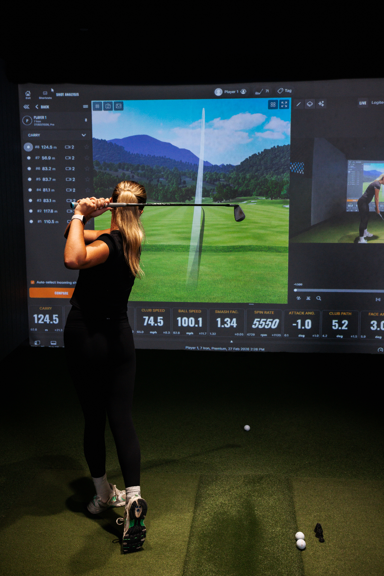 Golfer with iron play on the course simulator, carry and total data visible. Real course play, happening indoors.