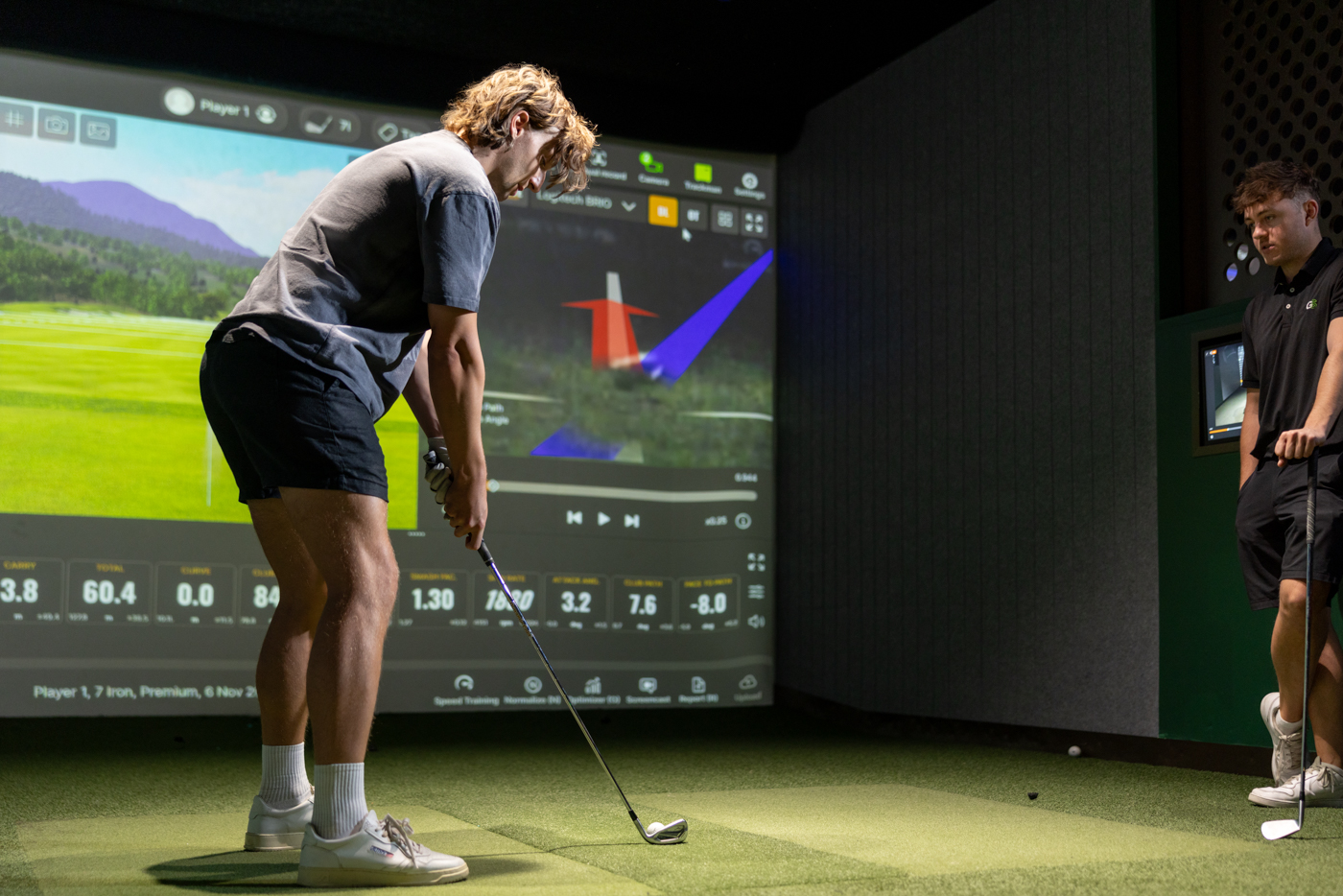 Golfer hitting an iron with a PGA coach watching and shot data on screen.